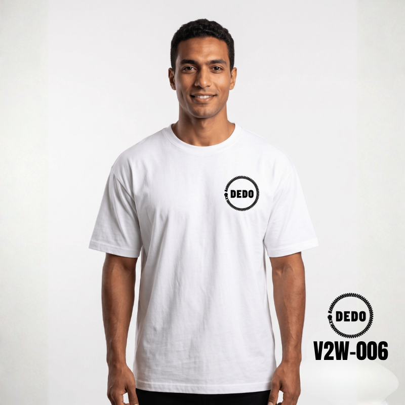 Rear view of a man walking down a historic cobblestone alleyway, wearing a white oversized t-shirt with a black futuristic cybernetic skull print on the back. The DEDO logo and product code V2W-006 are displayed in the bottom-left corner.