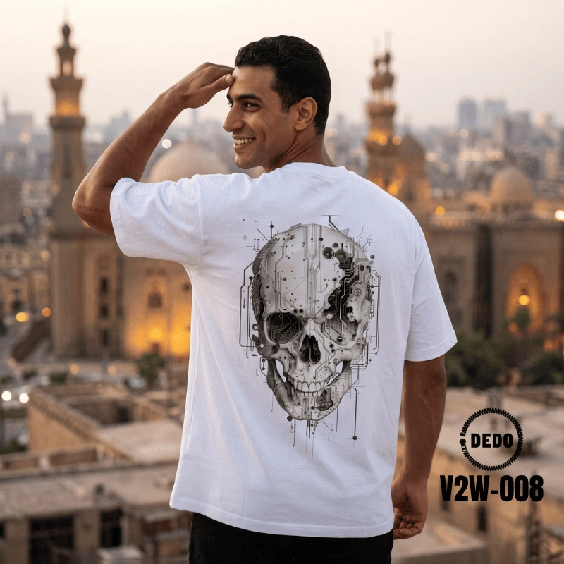 Rear view of a smiling man looking over his shoulder on a sunny waterfront promenade with feluccas in the background, wearing a white oversized t-shirt with a highly detailed, black cybernetic skull graphic print on the back integrated with circuit board wiring and mechanical details. The DEDO logo and product code V2W-008 are visible in the lower corner.